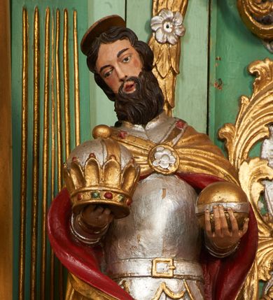 Zdjęcie nr 1: Święty ukazany frontalnie, z głową lekko przechyloną w prawo, w kontrapoście. Obie ręce ma ugięte, wyciągnięte przed siebie, w lewej dłoni trzyma jabłko królewskie, w prawej zamkniętą koronę. Twarz ma pociągłą, o wąskich oczach, prostym nosie i małych, otwartych ustach, okoloną gęstą brodą i ciemnymi, krótkimi włosami. Nad głową ma nimb. Ubrany jest w srebrzoną zbroję, ze złoconymi detalami oraz złocony płaszcz z czerwoną podszewką, zapięty na piersi ozdobną klamrą. Polichromia ciała naturalistyczna. 
