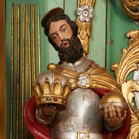 Zdjęcie nr 1: Święty ukazany frontalnie, z głową lekko przechyloną w prawo, w kontrapoście. Obie ręce ma ugięte, wyciągnięte przed siebie, w lewej dłoni trzyma jabłko królewskie, w prawej zamkniętą koronę. Twarz ma pociągłą, o wąskich oczach, prostym nosie i małych, otwartych ustach, okoloną gęstą brodą i ciemnymi, krótkimi włosami. Nad głową ma nimb. Ubrany jest w srebrzoną zbroję, ze złoconymi detalami oraz złocony płaszcz z czerwoną podszewką, zapięty na piersi ozdobną klamrą. Polichromia ciała naturalistyczna. 
