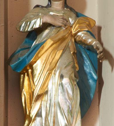 Zdjęcie nr 2: Rzeźba pełnoplastyczna, ustawiona na podstawie na planie kwadratu. Święta ukazana frontalnie, w kontrapoście, prawą rękę kładzie na piersi, lewą wyciąga w przód. Twarz ma pełną, o migdałowatych oczach, małym nosie i otwartych ustach, okoloną długimi, brązowymi włosami, opadającymi na ramiona. Na głowie ma koronę otwartą. Ubrana jest w srebrzoną suknię oraz złocony płaszcz z niebieską podszewką przewiązany diagonalnie przez lewe ramię. Polichromia w partiach ciała naturalistyczna. 
