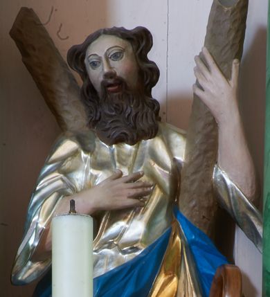 Zdjęcie nr 1: Rzeźba pełna, ustawiona na podstawie na planie kwadratu. Święty zwrócony frontalnie, z głową lekko skierowaną w prawo, w kontrapoście. Prawą rękę kładzie na piersi, lewą ujmuje ramię krzyża w kształcie litery „X”, stojącego za nim. Twarz ma pociągłą, o pełnych policzkach, dużych, migdałowatych oczach, prostym nosie i pełnych, rozchylonych ustach, okoloną brodą opadającą na pierś i falowanymi, brązowymi włosami sięgającymi ramion. Ubrany jest w srebrzoną, luźną tunikę oraz złocony płaszcz z niebieską podszewką zawiązany w partii nóg. Polichromia ciała naturalistyczna. 


