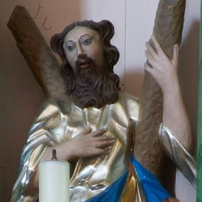 Zdjęcie nr 1: Rzeźba pełna, ustawiona na podstawie na planie kwadratu. Święty zwrócony frontalnie, z głową lekko skierowaną w prawo, w kontrapoście. Prawą rękę kładzie na piersi, lewą ujmuje ramię krzyża w kształcie litery „X”, stojącego za nim. Twarz ma pociągłą, o pełnych policzkach, dużych, migdałowatych oczach, prostym nosie i pełnych, rozchylonych ustach, okoloną brodą opadającą na pierś i falowanymi, brązowymi włosami sięgającymi ramion. Ubrany jest w srebrzoną, luźną tunikę oraz złocony płaszcz z niebieską podszewką zawiązany w partii nóg. Polichromia ciała naturalistyczna. 


