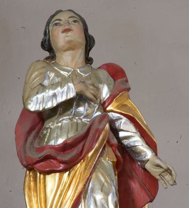 Zdjęcie nr 1: Rzeźba ścięta z tyłu, ustawiona na niskiej podstawie. Święty ukazany frontalnie, z uniesioną głową, prawą rękę kładzie na piersi, lewą, opuszczoną, wyciąga w bok. Twarz ma kwadratową, o wydatnym podbródku, wąskich oczach, małym nosie i ustach, okoloną ciemnymi, falowanymi włosami sięgającymi ramion. Ubrany jest srebrzoną tunikę z długimi rękawami i kołnierzykiem, przepasaną w talii oraz złoty płaszcz z czerwoną podszewką przewiązany diagonalnie przez lewe ramię. Polichromia w partiach ciała naturalistyczna.  