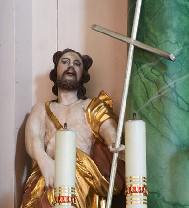 Zdjęcie nr 1: Rzeźba ścięta z tyłu, ustawiona na podstawie na planie kwadratu. Święty ukazany frontalnie, w kontrapoście. W lewej, wyciągniętej do przodu ręce trzyma laskę zakończoną krzyżem, prawą podtrzymuje z przodu płaszcz. Twarz ma owalną, o dużych, migdałowatych oczach, zadartym nosie i wąskich, otwartych ustach, okoloną krótką, silnie kręconą brodą oraz falowanymi włosami sięgającymi ramion. Ubrany jest w melotę zarzuconą na prawe ramię i zawiniętą wokół bioder, odsłaniającą klatkę piersiową i lewą nogę. Przy lewej nodze świętego srebrzony baranek. Polichromia ciała naturalistyczna. 

