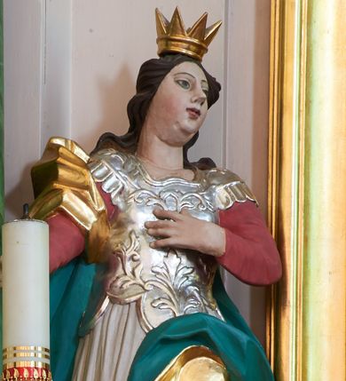 Zdjęcie nr 1: Rzeźba pełna, ustawiona na podstawie na planie kwadratu. Święta ukazana frontalnie, w kontrapoście, lewą rękę kładzie na piersi, prawą wyciąga w bok. Twarz ma pełną, o migdałowatych oczach, zadartym nosie i pełnych ustach, okoloną falowanymi, brązowymi włosami opadającymi na ramiona. Na głowie ma koronę otwartą. Święta ubrana jest w czerwono-różową suknię z długimi rękawami i sięgającą kolan srebrzoną suknię wierzchnią z gorsetem z płaskorzeźbioną dekoracją roślinną oraz pozłacany płaszcz z zieloną podszewką przerzucony przez prawe ramię, opadający na lewy bok. U stóp świętej fragment koła. Polichromia naturalistyczna w partiach ciała.
