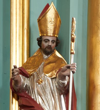 Zdjęcie nr 1: Rzeźba pełna, ustawiona na niskiej podstawie. Święty ukazany frontalnie, w kontrapoście, o esowato wygiętej sylwetce, obie ręce ma ugięte, wyciągnięte do przodu, prawa dłoń w geście błogosławieństwa, w drugiej trzyma pastorał. Twarz ma kwadratową, o migdałowatych oczach, małym nosie i pełnych ustach, okoloną krótką, brązową brodą oraz kręconymi włosami. Ubrany jest w długą, granatową albę, srebrzoną rokietę, na piersi ma zawieszony pektorał, a na ramiona narzuconą złoconą kapę z czerwoną podszewką, zapiętą na piersi klamrą, zawiniętą na prawej nodze. Na głowie ma złoconą infułę. Przy lewej nodze świętego niewielka postać klęczącego na lewe kolano Piotrowina z dłońmi złożonymi w geście modlitwy. Polichromia ciała naturalistyczna.