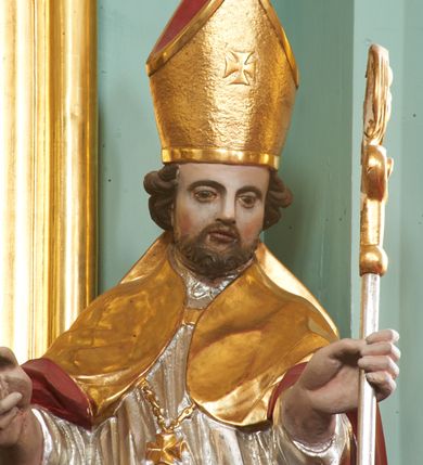 Zdjęcie nr 2: Rzeźba pełna, ustawiona na niskiej podstawie. Święty ukazany frontalnie, w kontrapoście, o esowato wygiętej sylwetce, obie ręce ma ugięte, wyciągnięte do przodu, prawa dłoń w geście błogosławieństwa, w drugiej trzyma pastorał. Twarz ma kwadratową, o migdałowatych oczach, małym nosie i pełnych ustach, okoloną krótką, brązową brodą oraz kręconymi włosami. Ubrany jest w długą, granatową albę, srebrzoną rokietę, na piersi ma zawieszony pektorał, a na ramiona narzuconą złoconą kapę z czerwoną podszewką, zapiętą na piersi klamrą, zawiniętą na prawej nodze. Na głowie ma złoconą infułę. Przy lewej nodze świętego niewielka postać klęczącego na lewe kolano Piotrowina z dłońmi złożonymi w geście modlitwy. Polichromia ciała naturalistyczna.