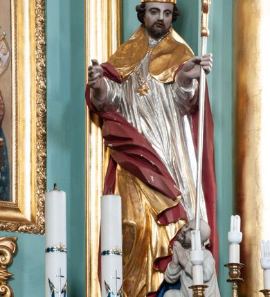 Zdjęcie nr 3: Rzeźba pełna, ustawiona na niskiej podstawie. Święty ukazany frontalnie, w kontrapoście, o esowato wygiętej sylwetce, obie ręce ma ugięte, wyciągnięte do przodu, prawa dłoń w geście błogosławieństwa, w drugiej trzyma pastorał. Twarz ma kwadratową, o migdałowatych oczach, małym nosie i pełnych ustach, okoloną krótką, brązową brodą oraz kręconymi włosami. Ubrany jest w długą, granatową albę, srebrzoną rokietę, na piersi ma zawieszony pektorał, a na ramiona narzuconą złoconą kapę z czerwoną podszewką, zapiętą na piersi klamrą, zawiniętą na prawej nodze. Na głowie ma złoconą infułę. Przy lewej nodze świętego niewielka postać klęczącego na lewe kolano Piotrowina z dłońmi złożonymi w geście modlitwy. Polichromia ciała naturalistyczna.