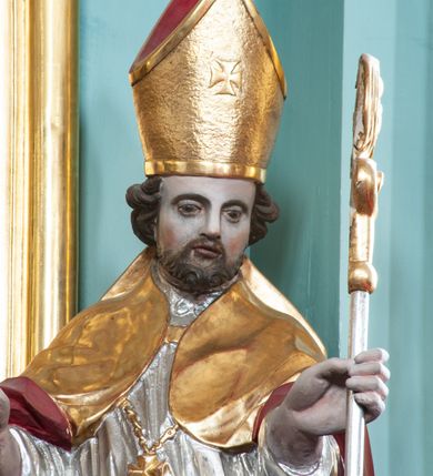 Zdjęcie nr 4: Rzeźba pełna, ustawiona na niskiej podstawie. Święty ukazany frontalnie, w kontrapoście, o esowato wygiętej sylwetce, obie ręce ma ugięte, wyciągnięte do przodu, prawa dłoń w geście błogosławieństwa, w drugiej trzyma pastorał. Twarz ma kwadratową, o migdałowatych oczach, małym nosie i pełnych ustach, okoloną krótką, brązową brodą oraz kręconymi włosami. Ubrany jest w długą, granatową albę, srebrzoną rokietę, na piersi ma zawieszony pektorał, a na ramiona narzuconą złoconą kapę z czerwoną podszewką, zapiętą na piersi klamrą, zawiniętą na prawej nodze. Na głowie ma złoconą infułę. Przy lewej nodze świętego niewielka postać klęczącego na lewe kolano Piotrowina z dłońmi złożonymi w geście modlitwy. Polichromia ciała naturalistyczna.