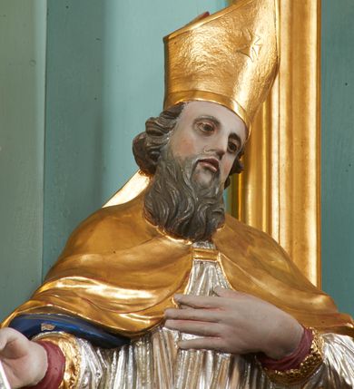 Zdjęcie nr 2: Rzeźba pełna, ustawiona na nieregularnej podstawie. Święty ukazany frontalnie, z głową przechyloną w lewo, w kontrapoście, o delikatnie przegiętej sylwetce. Lewą rękę ma ugiętą, uniesioną na wysokość piersi, w prawej trzyma laskę zakończoną krzyżem. Twarz ma pociągłą, o migdałowatych oczach, małym nosie i pełnych utach, okoloną brązową brodą i skręconymi, krótkimi włosami. Postać ubrana jest w długą, czerwoną albę, srebrzoną rokietę ze złoconym brzegiem oraz złoconą kapę z granatową podszewką, zapiętą klamrą na piersi i zawiniętą na lewej nodze. Na głowie ma złoconą infułę. Polichromia naturalistyczna w partiach ciała.