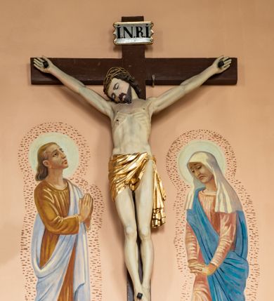 Zdjęcie nr 1: Rzeźba Chrystusa Ukrzyżowanego  między polichromowanymi na ścianie kościoła postaciami św. Jana Ewangelisty z lewej i Marii z prawej strony. Chrystus ukazany w lekkim zwisie, z wyprężonym ciałem, przybity do krzyża trzema gwoździami. Głowa opada mu na prawe ramię, oczy ma zamknięte, nos wąski, usta niewielki. Włosy opadają mu na plecy, ma krótkie wąsy i brodę. Jest przepasany  złotym perizonium. Nad głową ma titulus z napisem „INRI” pod stopami z napisem „Pamiątka ze starego kościoła”. Polichromia w partiach ciała naturalistyczna. 
Po lewej stronie krzyża młodzieńcze przedstawienie Jana Ewangelisty w  żółtej tunice i błękitnym płaszczu. Jest zwrócony w trzech czwartych w lewo, głowę kieruje w stronę Chrystusa, ręce ma złożone do modlitwy. Ma owalną twarz, ciemne oczy, wąski nos i  mięsiste usta, ciemnoblond włosy opadają my na kark. Matka Boska stoi po prawej stronie, zwrócona w stronę krzyża, ze spuszczoną głową; załamuje ręce. Ma smutną w wyrazie twarz, zamknięte oczy, duży nos i szerokie usta. Jest ubrana w różową suknię i błękitny płaszcz, na głowę ma zarzuconą białą chustę. Wokół głów postaci jasne nimby, ich ciała otoczone kreskowaniem, pod stopami zaznaczona linia ziemi. 
