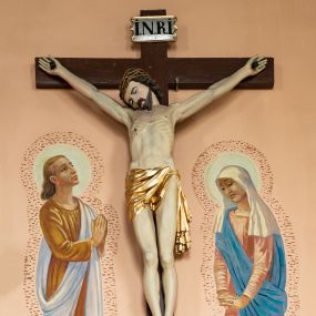 Zdjęcie nr 1: Rzeźba Chrystusa Ukrzyżowanego  między polichromowanymi na ścianie kościoła postaciami św. Jana Ewangelisty z lewej i Marii z prawej strony. Chrystus ukazany w lekkim zwisie, z wyprężonym ciałem, przybity do krzyża trzema gwoździami. Głowa opada mu na prawe ramię, oczy ma zamknięte, nos wąski, usta niewielki. Włosy opadają mu na plecy, ma krótkie wąsy i brodę. Jest przepasany  złotym perizonium. Nad głową ma titulus z napisem „INRI” pod stopami z napisem „Pamiątka ze starego kościoła”. Polichromia w partiach ciała naturalistyczna. 
Po lewej stronie krzyża młodzieńcze przedstawienie Jana Ewangelisty w  żółtej tunice i błękitnym płaszczu. Jest zwrócony w trzech czwartych w lewo, głowę kieruje w stronę Chrystusa, ręce ma złożone do modlitwy. Ma owalną twarz, ciemne oczy, wąski nos i  mięsiste usta, ciemnoblond włosy opadają my na kark. Matka Boska stoi po prawej stronie, zwrócona w stronę krzyża, ze spuszczoną głową; załamuje ręce. Ma smutną w wyrazie twarz, zamknięte oczy, duży nos i szerokie usta. Jest ubrana w różową suknię i błękitny płaszcz, na głowę ma zarzuconą białą chustę. Wokół głów postaci jasne nimby, ich ciała otoczone kreskowaniem, pod stopami zaznaczona linia ziemi. 
