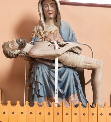 Zdjęcie nr 2: Matka Boska siedząca na tronie z płaskorzeźbioną dekoracją w formie maswerku, na jej kolanach horyzontalnie ułożone, martwe ciało Chrystusa. Dojrzała kobieta w bladoróżowej sukni i błękitnym płaszczu układającym się po bokach w kaskady fał. Poomiędzy nimi szaty uformowane w kształt litery V, na głowie biała chusta wykończona złotą lamówką. Ciało Chrystusa wychudzone z zaznaczonymi śladami męki, głowa w koronie cierniowej lekko odchylona do tyłu, włosy układające się w spiralne loki. Ręce złożone na wysokości bioder. Biodra ma owiniete jasnym perizonium.
