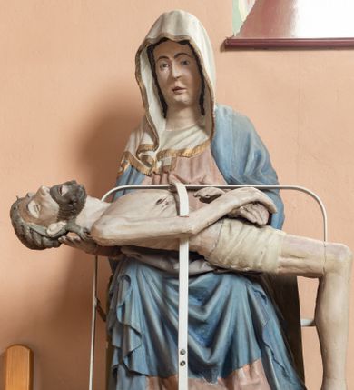 Zdjęcie nr 1: Matka Boska siedząca na tronie z płaskorzeźbioną dekoracją w formie maswerku, na jej kolanach horyzontalnie ułożone, martwe ciało Chrystusa. Dojrzała kobieta w bladoróżowej sukni i błękitnym płaszczu układającym się po bokach w kaskady fał. Poomiędzy nimi szaty uformowane w kształt litery V, na głowie biała chusta wykończona złotą lamówką. Ciało Chrystusa wychudzone z zaznaczonymi śladami męki, głowa w koronie cierniowej lekko odchylona do tyłu, włosy układające się w spiralne loki. Ręce złożone na wysokości bioder. Biodra ma owiniete jasnym perizonium.