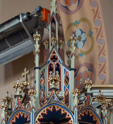 Zdjęcie nr 6: Ambona wisząca, przyścienna składająca się z kosza, zaplecka, baldachimu i schodów z balustradą. Kosz na planie wieloboku, podzielony na pola kolumienkami podtrzymującymi belkowanie, którego gzyms stanowi parapet. W polach płyciny w formie stojącego prostokąta zamkniętego łukiem ostrym. W płycinach całopostaciowe figury ewangelistów – od strony schodów – św. Jan, św. Marek, św. Łukasz i św. Mateusz. Każdy z nich ubrany w długą suknię, z narzuconym płaszczem, trzyma w rękach księgę lub zwój i ma (prócz Łukasza) przy nodze właściwy atrybut. Rzeźby są wolnostojące, na cokołach z podpisami. Wzdłuż ramy dolnej ażurowy ornament maswerkowy. Podstawa zwęża się ku dołowi, ścięta płasko. Zaplecek w kształcie stojącego prostokąta, wypełniony płyciną zamkniętą łukiem ostrym, ze zdwojonym łukiem i rozetą z czworoliściem.  Rozbudowany baldachim powtarza kształt kosza, składa się z arkad ujmujących zwieńczenie w formie kapliczki. Arkady od wewnątrz zdobi maswerkowy fryz, a wieńczą trójkątne, ażurowe szczyty z żabkami i kwiatonami. Wieńcząca kapliczka dwukondygnacyjna, z pinaklami, żabkami i kwiatonami; najwyższe pinakiel zakończony krzyżem. Struktura jasnozielona, niektóre elementy niebieskie lub czerwone, profile i ornamenty złocone.