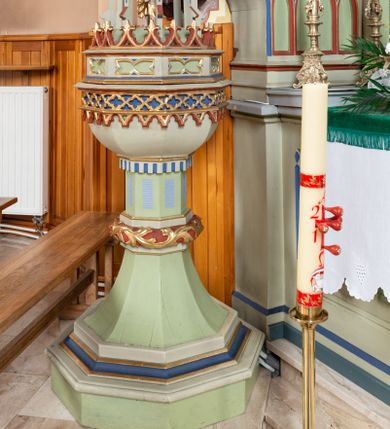 Zdjęcie nr 1: Chrzcielnica w typie kielichowym. Stopa na rzucie ośmioboku, na cokole, z uskokami, przechodzi w trzon, który w połowie wysokości przerwany jest spłaszczonym nodusem oplecionym suchą wicią akantową. Górna część trzonu z płycinami z karbowaniem. Czara sześcioboczna, ujęta jest dołem odwróconym krenelażem. Górna krawędź w formie wysuniętego gzymsu, poniżej ujęty dwoma wałkami rząd czworoliścia, z podwieszonymi, maswerkowymi, trójlistnymi arkadkami. Pokrywa w formie wieżyczki wypełnionej rzeźbiarską sceną Chrztu Chrystusa. Cokół dekorowany płycinami z leżącymi prostokątami o wklęsłych bokach, na nim fryz zakończony liliami. Ze środka sterczy dwukondygnacyjna, ażurowa wieżyczka. Każda kondygnacja skłąda sie z wspartych na filarach wimperg wypełnionych maswerkowaniem. Całość została zakończona pinaklem z żabkami i krzyżem. Między podporami pierwszej kondygnacji scena z klęczącym, składającym ręce na piersi Chrystusem i stojącym ponad nim Janem Chrzcicielem trzymającym drzewce w lewej ręce, a w prawej naczynie nad głową Zbawiciela. Struktura chrzcielnicy i pokrywy jasnozielona, niektóre elementy niebieskie lub czerwone, profile i ornamenty złocone. 