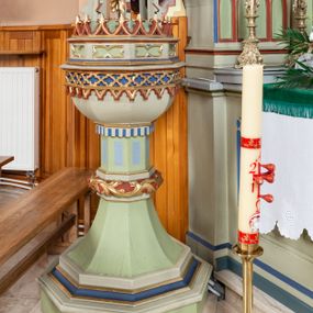 Zdjęcie nr 1: Chrzcielnica w typie kielichowym. Stopa na rzucie ośmioboku, na cokole, z uskokami, przechodzi w trzon, który w połowie wysokości przerwany jest spłaszczonym nodusem oplecionym suchą wicią akantową. Górna część trzonu z płycinami z karbowaniem. Czara sześcioboczna, ujęta jest dołem odwróconym krenelażem. Górna krawędź w formie wysuniętego gzymsu, poniżej ujęty dwoma wałkami rząd czworoliścia, z podwieszonymi, maswerkowymi, trójlistnymi arkadkami. Pokrywa w formie wieżyczki wypełnionej rzeźbiarską sceną Chrztu Chrystusa. Cokół dekorowany płycinami z leżącymi prostokątami o wklęsłych bokach, na nim fryz zakończony liliami. Ze środka sterczy dwukondygnacyjna, ażurowa wieżyczka. Każda kondygnacja skłąda sie z wspartych na filarach wimperg wypełnionych maswerkowaniem. Całość została zakończona pinaklem z żabkami i krzyżem. Między podporami pierwszej kondygnacji scena z klęczącym, składającym ręce na piersi Chrystusem i stojącym ponad nim Janem Chrzcicielem trzymającym drzewce w lewej ręce, a w prawej naczynie nad głową Zbawiciela. Struktura chrzcielnicy i pokrywy jasnozielona, niektóre elementy niebieskie lub czerwone, profile i ornamenty złocone. 