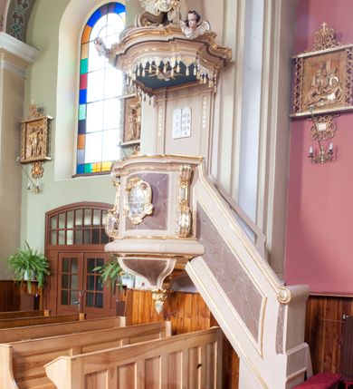 Zdjęcie nr 1: Ambona przyścienna, wisząca, dostępna schodami z podestem. Ich balustrada jest pełna, o poręczy w formie woluty, dekorowana płyciną. Kosz na profilowanym cokole; na planie prostokąta o wypukłej ściance frontowej i wklęsłej bocznej, wydzielonymi dwoma spływami wolutowymi, z płycinami. Od frontu kartusz dekorowany fragmentami rocaille’a i kampanulami z czernioną inskrypcją „KTO Z BOGA / JEST, SŁÓW / BOŻYCH SŁUCHA / JAN VIII”. Parapet w postaci profilowanego gzymsu wyłamanego nad wolutami. Spód kosza w kształcie wklęsło-wypukłego, odwróconego stożka zakończonego szyszką utworzoną z winnego grona otoczonego różami. Zaplecek w formie wydłużonego prostokąta ujętego pilastrami o trzonach dekorowanych płycinami i malowanymi złotem kampanulami. W polu płaskorzeźbione tablice Dekalogu. Baldachim powtarza plan kosza, składa się w podstawy utworzonej z profilowanego gzymsu z podwieszonym lambrekinem, na której symetrycznie ustawiono pięć schodzących się do środka wolut podtrzymujących cokół z figurą Chrystusa Dobrego Pasterza. Na gzymsie dwie uskrzydlone główki anielskie; w podniebiu baldachimu rzeźbiona gołębica Ducha Świętego w glorii, na niebieskim tle. Struktura w odcieniu bladego różu, płyciny marmoryzowane; dekoracja i profile złocone i srebrzone. Polichromia w odcieniu bladego różu, ścianki marmoryzowane; dekoracja i profile złocone i srebrzone.