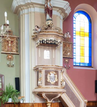 Zdjęcie nr 2: Ambona przyścienna, wisząca, dostępna schodami z podestem. Ich balustrada jest pełna, o poręczy w formie woluty, dekorowana płyciną. Kosz na profilowanym cokole; na planie prostokąta o wypukłej ściance frontowej i wklęsłej bocznej, wydzielonymi dwoma spływami wolutowymi, z płycinami. Od frontu kartusz dekorowany fragmentami rocaille’a i kampanulami z czernioną inskrypcją „KTO Z BOGA / JEST, SŁÓW / BOŻYCH SŁUCHA / JAN VIII”. Parapet w postaci profilowanego gzymsu wyłamanego nad wolutami. Spód kosza w kształcie wklęsło-wypukłego, odwróconego stożka zakończonego szyszką utworzoną z winnego grona otoczonego różami. Zaplecek w formie wydłużonego prostokąta ujętego pilastrami o trzonach dekorowanych płycinami i malowanymi złotem kampanulami. W polu płaskorzeźbione tablice Dekalogu. Baldachim powtarza plan kosza, składa się w podstawy utworzonej z profilowanego gzymsu z podwieszonym lambrekinem, na której symetrycznie ustawiono pięć schodzących się do środka wolut podtrzymujących cokół z figurą Chrystusa Dobrego Pasterza. Na gzymsie dwie uskrzydlone główki anielskie; w podniebiu baldachimu rzeźbiona gołębica Ducha Świętego w glorii, na niebieskim tle. Struktura w odcieniu bladego różu, płyciny marmoryzowane; dekoracja i profile złocone i srebrzone. Polichromia w odcieniu bladego różu, ścianki marmoryzowane; dekoracja i profile złocone i srebrzone.