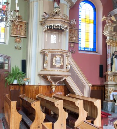 Zdjęcie nr 3: Ambona przyścienna, wisząca, dostępna schodami z podestem. Ich balustrada jest pełna, o poręczy w formie woluty, dekorowana płyciną. Kosz na profilowanym cokole; na planie prostokąta o wypukłej ściance frontowej i wklęsłej bocznej, wydzielonymi dwoma spływami wolutowymi, z płycinami. Od frontu kartusz dekorowany fragmentami rocaille’a i kampanulami z czernioną inskrypcją „KTO Z BOGA / JEST, SŁÓW / BOŻYCH SŁUCHA / JAN VIII”. Parapet w postaci profilowanego gzymsu wyłamanego nad wolutami. Spód kosza w kształcie wklęsło-wypukłego, odwróconego stożka zakończonego szyszką utworzoną z winnego grona otoczonego różami. Zaplecek w formie wydłużonego prostokąta ujętego pilastrami o trzonach dekorowanych płycinami i malowanymi złotem kampanulami. W polu płaskorzeźbione tablice Dekalogu. Baldachim powtarza plan kosza, składa się w podstawy utworzonej z profilowanego gzymsu z podwieszonym lambrekinem, na której symetrycznie ustawiono pięć schodzących się do środka wolut podtrzymujących cokół z figurą Chrystusa Dobrego Pasterza. Na gzymsie dwie uskrzydlone główki anielskie; w podniebiu baldachimu rzeźbiona gołębica Ducha Świętego w glorii, na niebieskim tle. Struktura w odcieniu bladego różu, płyciny marmoryzowane; dekoracja i profile złocone i srebrzone. Polichromia w odcieniu bladego różu, ścianki marmoryzowane; dekoracja i profile złocone i srebrzone.