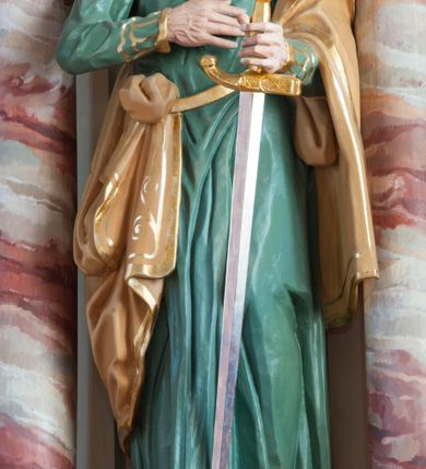 Zdjęcie nr 2: Rzeźba pełna, ustawiona na prostopadłościennym cokole. Figura ukazana w całej postaci, zwrócona frontalnie, w delikatnym kontrapoście, z lewym kolanem zgiętym. Głowa ustawiona na wprost, twarz podłużna, szczupła, okolona gęstym zarostem; wzrok zwrócony przed siebie. Ciemne włosy formują lok nad czołem i opadają na kark zasłaniając uszy, wąsy są szerokie, broda dzieli się w dwa pukle. Święty prawą, zgiętą rękę przyciska do piersi, w wyciągniętej przed siebie lewej trzyma rękojeść opartego o ziemię miecza. Apostoł jest ubrany zieloną w suknię sięgającą ziemi, wykończoną przy krawędziach złotym ornamentem geometrycznym; tkanina łamie się w sztywne, drobne fałdki. Beżowy, dekorowany złotym ornamentem roślinnym płaszcz otacza lewą rękę i jest zatknięty za pasem przy prawym biodrze. Polichromia naturalistyczna w odsłoniętych partiach ciała, miecz złocony i srebrzony. 
