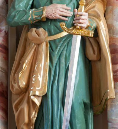 Zdjęcie nr 3: Rzeźba pełna, ustawiona na prostopadłościennym cokole. Figura ukazana w całej postaci, zwrócona frontalnie, w delikatnym kontrapoście, z lewym kolanem zgiętym. Głowa ustawiona na wprost, twarz podłużna, szczupła, okolona gęstym zarostem; wzrok zwrócony przed siebie. Ciemne włosy formują lok nad czołem i opadają na kark zasłaniając uszy, wąsy są szerokie, broda dzieli się w dwa pukle. Święty prawą, zgiętą rękę przyciska do piersi, w wyciągniętej przed siebie lewej trzyma rękojeść opartego o ziemię miecza. Apostoł jest ubrany zieloną w suknię sięgającą ziemi, wykończoną przy krawędziach złotym ornamentem geometrycznym; tkanina łamie się w sztywne, drobne fałdki. Beżowy, dekorowany złotym ornamentem roślinnym płaszcz otacza lewą rękę i jest zatknięty za pasem przy prawym biodrze. Polichromia naturalistyczna w odsłoniętych partiach ciała, miecz złocony i srebrzony. 