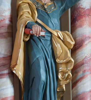 Zdjęcie nr 2: Rzeźba pełna, ustawiona na prostopadłościennym cokole. Figura ukazana w całej postaci, zwrócona frontalnie, w kontrapoście, z prawą nogą zgiętą. Głowa przechylona na lewe ramię, uniesiona; twarz prostokątna, okolona zarostem; wzrok uniesiony. Siwiejące włosy formują lok nad czołem, wąsy dzielą się na równoległe pasemka, broda skręcona w niewielkie loki. Święty prawą ręką dociska do ciała zamkniętą księgę, w lewej, uniesionej trzyma dwa klucze. Apostoł jest ubrany w niebieską suknię sięgającą ziemi, wykończoną przy krawędziach złotym ornamentem; beżowy, lamowany złotem płaszcz otacza prawą rękę i jest zatknięty za pasem przy lewym biodrze, opada ku ziemi dekoracyjnymi fałdami. Polichromia naturalistyczna w odsłoniętych partiach ciała, księga czerwona, o białych kartach, jeden klucz złocony, drugi srebrzony. 
