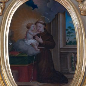 Zdjęcie nr 1: Obraz w kształcie stojącego owalu z przedstawieniem postaci św. Antoniego ujęty profilowaną, złoconą ramą. Święty ukazany został całopostaciowo, w trzech czwartych, zwrócony w prawo. Klęczy przed stołem, na którym siedzi przytrzymywane przez niego Dzieciątko Jezus. Ma delikatną, młodzieńczą twarz, półprzymknięte oczy, wąski nos i usta, a na głowie wyciętą tonsurę, ponad nią nimb. Jest ubrany w brązowy habit franciszkański przewiązany sznurem. Lewą rękę przyciska do piersi. Dzieciątko zostało ukazane z profilu, siedząc na szarej chmurze, z biodrami osłoniętymi białą szarfą. Obiema rękami ujmuje świętego za szyję, głowę rozświetla mu szeroki, promienisty nimb, wokół rozchodzą się obłoki. Poniżej chmury, przy krawędzi kompozycji widoczne leżące na okrytym zieloną tkaniną ze złotym obszyciem blacie księga i lilia. Scena umieszczona jest we wnętrzu lub na balkonie otwierającym się prostokątnym oknem na pejzaż, poniżej widoczny fragment balustrady. Kolorystyka nasycona, ciepła. 