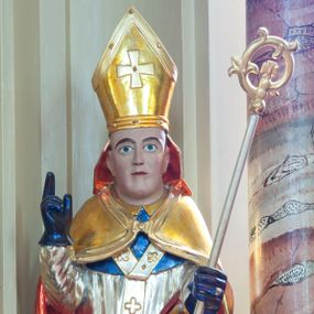 Zdjęcie nr 1: Figura drążona, na cokole. Święty Stanisław przedstawiony w całej postaci, stojący frontalnie, obie ręce ma zgięte w łokciach, prawą unosi do góry, lewą przytrzymuje pastorał. Stanisław ma pociągła twarz z dużymi, szeroko otwartymi oczami, szerokim nosem i wąskimi ustami. Jest ubrany w strój biskupi: niebieską albę, białą rokietę zdobioną złotym pasem koronki przy dolnej krawędzi i paliusz z aplikowanymi krzyżami greckimi. Na to założoną ma obszerną, złotą kapę podbitą czerwienią, która spięta pod szyją okrywa ramiona i spływa do ziemi; na głowie ma infułę, a na rękach niebieskie rękawiczki. Polichromia w odkrytych partiach karnacji postaci jest traktowana naturalistycznie.