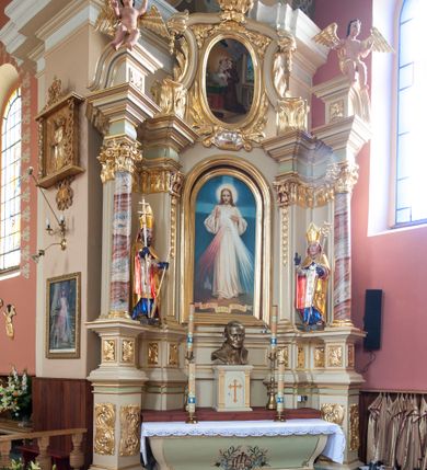 Zdjęcie nr 2: Figura drążona, na cokole. Święty Stanisław przedstawiony w całej postaci, stojący frontalnie, obie ręce ma zgięte w łokciach, prawą unosi do góry, lewą przytrzymuje pastorał. Stanisław ma pociągła twarz z dużymi, szeroko otwartymi oczami, szerokim nosem i wąskimi ustami. Jest ubrany w strój biskupi: niebieską albę, białą rokietę zdobioną złotym pasem koronki przy dolnej krawędzi i paliusz z aplikowanymi krzyżami greckimi. Na to założoną ma obszerną, złotą kapę podbitą czerwienią, która spięta pod szyją okrywa ramiona i spływa do ziemi; na głowie ma infułę, a na rękach niebieskie rękawiczki. Polichromia w odkrytych partiach karnacji postaci jest traktowana naturalistycznie.