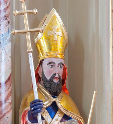 Zdjęcie nr 1: Figura drążona, na cokole. Święty Wojciech przedstawiony w całej postaci, stojący frontalnie, obie ręce ma zgięte w łokciach, w prawej trzyma ferulę, czyli pastorał zakończony podwójnym krzyżem, lewą przytrzymuje oparte o ziemię wiosło. Wojciech ma pociągła twarz z dużymi, szeroko otwartymi oczami, szerokim nosem i ustami o kącikach skierowanych w dół. Ciemne włosy ma zaczesane do tyłu, gęsta, długa broda równoległymi pasmami opada na pierś. Jest ubrany w strój biskupi: niebieską albę, białą rokietę zdobioną złotym pasem koronki przy dolnej krawędzi i paliusz z aplikowanymi krzyżami greckimi. Na to założoną ma obszerną, złotą kapę podbitą czerwienią, która spięta pod szyją okrywa ramiona i spływa do ziemi; na głowie ma infułę, a na rękach niebieskie rękawiczki. Polichromia w odkrytych partiach karnacji postaci jest traktowana naturalistycznie. 