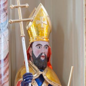 Zdjęcie nr 1: Figura drążona, na cokole. Święty Wojciech przedstawiony w całej postaci, stojący frontalnie, obie ręce ma zgięte w łokciach, w prawej trzyma ferulę, czyli pastorał zakończony podwójnym krzyżem, lewą przytrzymuje oparte o ziemię wiosło. Wojciech ma pociągła twarz z dużymi, szeroko otwartymi oczami, szerokim nosem i ustami o kącikach skierowanych w dół. Ciemne włosy ma zaczesane do tyłu, gęsta, długa broda równoległymi pasmami opada na pierś. Jest ubrany w strój biskupi: niebieską albę, białą rokietę zdobioną złotym pasem koronki przy dolnej krawędzi i paliusz z aplikowanymi krzyżami greckimi. Na to założoną ma obszerną, złotą kapę podbitą czerwienią, która spięta pod szyją okrywa ramiona i spływa do ziemi; na głowie ma infułę, a na rękach niebieskie rękawiczki. Polichromia w odkrytych partiach karnacji postaci jest traktowana naturalistycznie. 