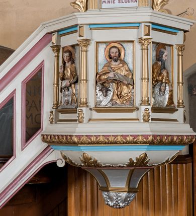 Zdjęcie nr 7: Ambona przyścienna, wisząca, z dostępem od lewej strony w postaci jednobiegowych schodów z balustradą. Kosz na planie sześcioboku na profilowanym cokole i ćwierćwałku; u spodu podwieszony profilowany ostrosłup o ściętym szczycie, zakończony szyszką. Ścianki kosza wydzielone kolumienkami  o trzonach dekorowanych do 1/3 wysokości kanelowaniem, o kapitelach korynckich, na cokołach zdobionych liśćmi akantu, dźwigającymi imposty oraz wspólny gzyms stanowiąc parapet. W polach ścianek płyciny z płaskorzeźbionymi półpostaciami czterech ewangelistów, każdy z nich ukazany został z złotej sukni, z identyfikującym go atrybutem, księgą lub zwojem i piórem w ręce –  od lewej św. Jan, św. Mateusz, św. Łukasz i św. Marek. Zaplecek w formie wydłużonego prostokąta, ujętego pilastrami podtrzymującymi imposty i wspólny gzyms. W polu płycina w formie stojącego prostokąta zamkniętego łukiem półkolistym z płaskorzeźbionym przedstawieniem Chrystusa Dobrego Pasterza, u spodu płycina z napisem „KTO WAS SŁUCHA / MNIE SŁUCHA”.  Chrystus został ukazany w całej postaci, lewą ręką podtrzymując baranka, a prawą laskę pasterską. Ma podłużna, poważną twarz, małe, niebieskie oczy, wąski nos i usta, broda dzieli się w dwa pukle; falowane włosy opadają na ramiona. Jest ubrany w długą, złotą tunikę i złoty płaszcz, na głowie ma złoty kalepusz, czerwony od wewnątrz. Stoi na trawie, u jego stóp dwa srebrzone baranki Zaplecek ujęty uszami w formie falujących liści. Baldachim powtarza kształt kosza, nakryty kopułką pokrytą łuską, z podwieszonym fryzem roślinnym, dekorowany w narożach palmetami. Baldachim stanowi podstawę dla latarni opartej na sześciu spływach wolutowych dekorowanych kampanulami, podtrzymujących belkowanie i kopułkę, stanowiącą cokół dla figury św. Jana Chrzciciela, który stoi w kontrapoście, prawą ręką unosi na wysokość twarzy muszlę, a e lewej trzyma trzyż. Ma podł€żną twarz okoczoną gęstym zarostem, wzniesiony ku górze wzrok, wąski nos i małe usta. Jest ubrany w tunikę ze srerzonego fuyta i złoty płaszcz wywiniety na prawym biodrze i przechodzący przez lewy bark. Wewnątrz latarni figura anioła na postumencie. Został ukazany w całej postaci, z rękami złożonymi na piersi. Ma okrągłą twarz o deliatnych rysach i falowane włosy opadające na kark. Jest ubrany w długa, złotą suknię w szerokomi rękawami. Z pleców wyrastają mu duże, srebrne skrzydła z ptacich piór. Na podniebiu baldachimu gołębica Ducha Świętego w glorii promienistej. Struktura polichromowana na biało, profile na czerwono, detale i ornamentyka złocone.