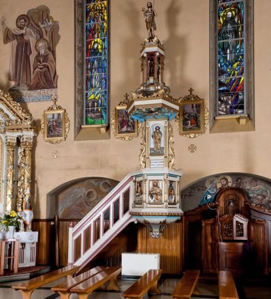 Zdjęcie nr 2: Ambona przyścienna, wisząca, z dostępem od lewej strony w postaci jednobiegowych schodów z balustradą. Kosz na planie sześcioboku na profilowanym cokole i ćwierćwałku; u spodu podwieszony profilowany ostrosłup o ściętym szczycie, zakończony szyszką. Ścianki kosza wydzielone kolumienkami  o trzonach dekorowanych do 1/3 wysokości kanelowaniem, o kapitelach korynckich, na cokołach zdobionych liśćmi akantu, dźwigającymi imposty oraz wspólny gzyms stanowiąc parapet. W polach ścianek płyciny z płaskorzeźbionymi półpostaciami czterech ewangelistów, każdy z nich ukazany został z złotej sukni, z identyfikującym go atrybutem, księgą lub zwojem i piórem w ręce –  od lewej św. Jan, św. Mateusz, św. Łukasz i św. Marek. Zaplecek w formie wydłużonego prostokąta, ujętego pilastrami podtrzymującymi imposty i wspólny gzyms. W polu płycina w formie stojącego prostokąta zamkniętego łukiem półkolistym z płaskorzeźbionym przedstawieniem Chrystusa Dobrego Pasterza, u spodu płycina z napisem „KTO WAS SŁUCHA / MNIE SŁUCHA”.  Chrystus został ukazany w całej postaci, lewą ręką podtrzymując baranka, a prawą laskę pasterską. Ma podłużna, poważną twarz, małe, niebieskie oczy, wąski nos i usta, broda dzieli się w dwa pukle; falowane włosy opadają na ramiona. Jest ubrany w długą, złotą tunikę i złoty płaszcz, na głowie ma złoty kalepusz, czerwony od wewnątrz. Stoi na trawie, u jego stóp dwa srebrzone baranki Zaplecek ujęty uszami w formie falujących liści. Baldachim powtarza kształt kosza, nakryty kopułką pokrytą łuską, z podwieszonym fryzem roślinnym, dekorowany w narożach palmetami. Baldachim stanowi podstawę dla latarni opartej na sześciu spływach wolutowych dekorowanych kampanulami, podtrzymujących belkowanie i kopułkę, stanowiącą cokół dla figury św. Jana Chrzciciela, który stoi w kontrapoście, prawą ręką unosi na wysokość twarzy muszlę, a e lewej trzyma trzyż. Ma podł€żną twarz okoczoną gęstym zarostem, wzniesiony ku górze wzrok, wąski nos i małe usta. Jest ubrany w tunikę ze srerzonego fuyta i złoty płaszcz wywiniety na prawym biodrze i przechodzący przez lewy bark. Wewnątrz latarni figura anioła na postumencie. Został ukazany w całej postaci, z rękami złożonymi na piersi. Ma okrągłą twarz o deliatnych rysach i falowane włosy opadające na kark. Jest ubrany w długa, złotą suknię w szerokomi rękawami. Z pleców wyrastają mu duże, srebrne skrzydła z ptacich piór. Na podniebiu baldachimu gołębica Ducha Świętego w glorii promienistej. Struktura polichromowana na biało, profile na czerwono, detale i ornamentyka złocone.