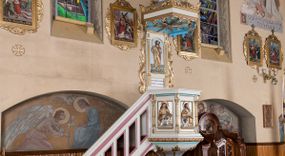 Zdjęcie nr 1: Ambona przyścienna, wisząca, z dostępem od lewej strony w postaci jednobiegowych schodów z balustradą. Kosz na planie sześcioboku na profilowanym cokole i ćwierćwałku; u spodu podwieszony profilowany ostrosłup o ściętym szczycie, zakończony szyszką. Ścianki kosza wydzielone kolumienkami  o trzonach dekorowanych do 1/3 wysokości kanelowaniem, o kapitelach korynckich, na cokołach zdobionych liśćmi akantu, dźwigającymi imposty oraz wspólny gzyms stanowiąc parapet. W polach ścianek płyciny z płaskorzeźbionymi półpostaciami czterech ewangelistów, każdy z nich ukazany został z złotej sukni, z identyfikującym go atrybutem, księgą lub zwojem i piórem w ręce –  od lewej św. Jan, św. Mateusz, św. Łukasz i św. Marek. Zaplecek w formie wydłużonego prostokąta, ujętego pilastrami podtrzymującymi imposty i wspólny gzyms. W polu płycina w formie stojącego prostokąta zamkniętego łukiem półkolistym z płaskorzeźbionym przedstawieniem Chrystusa Dobrego Pasterza, u spodu płycina z napisem „KTO WAS SŁUCHA / MNIE SŁUCHA”.  Chrystus został ukazany w całej postaci, lewą ręką podtrzymując baranka, a prawą laskę pasterską. Ma podłużna, poważną twarz, małe, niebieskie oczy, wąski nos i usta, broda dzieli się w dwa pukle; falowane włosy opadają na ramiona. Jest ubrany w długą, złotą tunikę i złoty płaszcz, na głowie ma złoty kalepusz, czerwony od wewnątrz. Stoi na trawie, u jego stóp dwa srebrzone baranki Zaplecek ujęty uszami w formie falujących liści. Baldachim powtarza kształt kosza, nakryty kopułką pokrytą łuską, z podwieszonym fryzem roślinnym, dekorowany w narożach palmetami. Baldachim stanowi podstawę dla latarni opartej na sześciu spływach wolutowych dekorowanych kampanulami, podtrzymujących belkowanie i kopułkę, stanowiącą cokół dla figury św. Jana Chrzciciela, który stoi w kontrapoście, prawą ręką unosi na wysokość twarzy muszlę, a e lewej trzyma trzyż. Ma podł€żną twarz okoczoną gęstym zarostem, wzniesiony ku górze wzrok, wąski nos i małe usta. Jest ubrany w tunikę ze srerzonego fuyta i złoty płaszcz wywiniety na prawym biodrze i przechodzący przez lewy bark. Wewnątrz latarni figura anioła na postumencie. Został ukazany w całej postaci, z rękami złożonymi na piersi. Ma okrągłą twarz o deliatnych rysach i falowane włosy opadające na kark. Jest ubrany w długa, złotą suknię w szerokomi rękawami. Z pleców wyrastają mu duże, srebrne skrzydła z ptacich piór. Na podniebiu baldachimu gołębica Ducha Świętego w glorii promienistej. Struktura polichromowana na biało, profile na czerwono, detale i ornamentyka złocone.