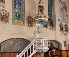 Zdjęcie nr 1: Ambona przyścienna, wisząca, z dostępem od lewej strony w postaci jednobiegowych schodów z balustradą. Kosz na planie sześcioboku na profilowanym cokole i ćwierćwałku; u spodu podwieszony profilowany ostrosłup o ściętym szczycie, zakończony szyszką. Ścianki kosza wydzielone kolumienkami  o trzonach dekorowanych do 1/3 wysokości kanelowaniem, o kapitelach korynckich, na cokołach zdobionych liśćmi akantu, dźwigającymi imposty oraz wspólny gzyms stanowiąc parapet. W polach ścianek płyciny z płaskorzeźbionymi półpostaciami czterech ewangelistów, każdy z nich ukazany został z złotej sukni, z identyfikującym go atrybutem, księgą lub zwojem i piórem w ręce –  od lewej św. Jan, św. Mateusz, św. Łukasz i św. Marek. Zaplecek w formie wydłużonego prostokąta, ujętego pilastrami podtrzymującymi imposty i wspólny gzyms. W polu płycina w formie stojącego prostokąta zamkniętego łukiem półkolistym z płaskorzeźbionym przedstawieniem Chrystusa Dobrego Pasterza, u spodu płycina z napisem „KTO WAS SŁUCHA / MNIE SŁUCHA”.  Chrystus został ukazany w całej postaci, lewą ręką podtrzymując baranka, a prawą laskę pasterską. Ma podłużna, poważną twarz, małe, niebieskie oczy, wąski nos i usta, broda dzieli się w dwa pukle; falowane włosy opadają na ramiona. Jest ubrany w długą, złotą tunikę i złoty płaszcz, na głowie ma złoty kalepusz, czerwony od wewnątrz. Stoi na trawie, u jego stóp dwa srebrzone baranki Zaplecek ujęty uszami w formie falujących liści. Baldachim powtarza kształt kosza, nakryty kopułką pokrytą łuską, z podwieszonym fryzem roślinnym, dekorowany w narożach palmetami. Baldachim stanowi podstawę dla latarni opartej na sześciu spływach wolutowych dekorowanych kampanulami, podtrzymujących belkowanie i kopułkę, stanowiącą cokół dla figury św. Jana Chrzciciela, który stoi w kontrapoście, prawą ręką unosi na wysokość twarzy muszlę, a e lewej trzyma trzyż. Ma podł€żną twarz okoczoną gęstym zarostem, wzniesiony ku górze wzrok, wąski nos i małe usta. Jest ubrany w tunikę ze srerzonego fuyta i złoty płaszcz wywiniety na prawym biodrze i przechodzący przez lewy bark. Wewnątrz latarni figura anioła na postumencie. Został ukazany w całej postaci, z rękami złożonymi na piersi. Ma okrągłą twarz o deliatnych rysach i falowane włosy opadające na kark. Jest ubrany w długa, złotą suknię w szerokomi rękawami. Z pleców wyrastają mu duże, srebrne skrzydła z ptacich piór. Na podniebiu baldachimu gołębica Ducha Świętego w glorii promienistej. Struktura polichromowana na biało, profile na czerwono, detale i ornamentyka złocone.