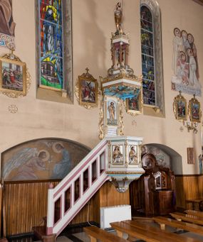 Zdjęcie nr 1: Ambona przyścienna, wisząca, z dostępem od lewej strony w postaci jednobiegowych schodów z balustradą. Kosz na planie sześcioboku na profilowanym cokole i ćwierćwałku; u spodu podwieszony profilowany ostrosłup o ściętym szczycie, zakończony szyszką. Ścianki kosza wydzielone kolumienkami  o trzonach dekorowanych do 1/3 wysokości kanelowaniem, o kapitelach korynckich, na cokołach zdobionych liśćmi akantu, dźwigającymi imposty oraz wspólny gzyms stanowiąc parapet. W polach ścianek płyciny z płaskorzeźbionymi półpostaciami czterech ewangelistów, każdy z nich ukazany został z złotej sukni, z identyfikującym go atrybutem, księgą lub zwojem i piórem w ręce –  od lewej św. Jan, św. Mateusz, św. Łukasz i św. Marek. Zaplecek w formie wydłużonego prostokąta, ujętego pilastrami podtrzymującymi imposty i wspólny gzyms. W polu płycina w formie stojącego prostokąta zamkniętego łukiem półkolistym z płaskorzeźbionym przedstawieniem Chrystusa Dobrego Pasterza, u spodu płycina z napisem „KTO WAS SŁUCHA / MNIE SŁUCHA”.  Chrystus został ukazany w całej postaci, lewą ręką podtrzymując baranka, a prawą laskę pasterską. Ma podłużna, poważną twarz, małe, niebieskie oczy, wąski nos i usta, broda dzieli się w dwa pukle; falowane włosy opadają na ramiona. Jest ubrany w długą, złotą tunikę i złoty płaszcz, na głowie ma złoty kalepusz, czerwony od wewnątrz. Stoi na trawie, u jego stóp dwa srebrzone baranki Zaplecek ujęty uszami w formie falujących liści. Baldachim powtarza kształt kosza, nakryty kopułką pokrytą łuską, z podwieszonym fryzem roślinnym, dekorowany w narożach palmetami. Baldachim stanowi podstawę dla latarni opartej na sześciu spływach wolutowych dekorowanych kampanulami, podtrzymujących belkowanie i kopułkę, stanowiącą cokół dla figury św. Jana Chrzciciela, który stoi w kontrapoście, prawą ręką unosi na wysokość twarzy muszlę, a e lewej trzyma trzyż. Ma podł€żną twarz okoczoną gęstym zarostem, wzniesiony ku górze wzrok, wąski nos i małe usta. Jest ubrany w tunikę ze srerzonego fuyta i złoty płaszcz wywiniety na prawym biodrze i przechodzący przez lewy bark. Wewnątrz latarni figura anioła na postumencie. Został ukazany w całej postaci, z rękami złożonymi na piersi. Ma okrągłą twarz o deliatnych rysach i falowane włosy opadające na kark. Jest ubrany w długa, złotą suknię w szerokomi rękawami. Z pleców wyrastają mu duże, srebrne skrzydła z ptacich piór. Na podniebiu baldachimu gołębica Ducha Świętego w glorii promienistej. Struktura polichromowana na biało, profile na czerwono, detale i ornamentyka złocone.