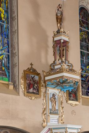 Zdjęcie nr 1: Ambona przyścienna, wisząca, z dostępem od lewej strony w postaci jednobiegowych schodów z balustradą. Kosz na planie sześcioboku na profilowanym cokole i ćwierćwałku; u spodu podwieszony profilowany ostrosłup o ściętym szczycie, zakończony szyszką. Ścianki kosza wydzielone kolumienkami  o trzonach dekorowanych do 1/3 wysokości kanelowaniem, o kapitelach korynckich, na cokołach zdobionych liśćmi akantu, dźwigającymi imposty oraz wspólny gzyms stanowiąc parapet. W polach ścianek płyciny z płaskorzeźbionymi półpostaciami czterech ewangelistów, każdy z nich ukazany został z złotej sukni, z identyfikującym go atrybutem, księgą lub zwojem i piórem w ręce –  od lewej św. Jan, św. Mateusz, św. Łukasz i św. Marek. Zaplecek w formie wydłużonego prostokąta, ujętego pilastrami podtrzymującymi imposty i wspólny gzyms. W polu płycina w formie stojącego prostokąta zamkniętego łukiem półkolistym z płaskorzeźbionym przedstawieniem Chrystusa Dobrego Pasterza, u spodu płycina z napisem „KTO WAS SŁUCHA / MNIE SŁUCHA”.  Chrystus został ukazany w całej postaci, lewą ręką podtrzymując baranka, a prawą laskę pasterską. Ma podłużna, poważną twarz, małe, niebieskie oczy, wąski nos i usta, broda dzieli się w dwa pukle; falowane włosy opadają na ramiona. Jest ubrany w długą, złotą tunikę i złoty płaszcz, na głowie ma złoty kalepusz, czerwony od wewnątrz. Stoi na trawie, u jego stóp dwa srebrzone baranki Zaplecek ujęty uszami w formie falujących liści. Baldachim powtarza kształt kosza, nakryty kopułką pokrytą łuską, z podwieszonym fryzem roślinnym, dekorowany w narożach palmetami. Baldachim stanowi podstawę dla latarni opartej na sześciu spływach wolutowych dekorowanych kampanulami, podtrzymujących belkowanie i kopułkę, stanowiącą cokół dla figury św. Jana Chrzciciela, który stoi w kontrapoście, prawą ręką unosi na wysokość twarzy muszlę, a e lewej trzyma trzyż. Ma podł€żną twarz okoczoną gęstym zarostem, wzniesiony ku górze wzrok, wąski nos i małe usta. Jest ubrany w tunikę ze srerzonego fuyta i złoty płaszcz wywiniety na prawym biodrze i przechodzący przez lewy bark. Wewnątrz latarni figura anioła na postumencie. Został ukazany w całej postaci, z rękami złożonymi na piersi. Ma okrągłą twarz o deliatnych rysach i falowane włosy opadające na kark. Jest ubrany w długa, złotą suknię w szerokomi rękawami. Z pleców wyrastają mu duże, srebrne skrzydła z ptacich piór. Na podniebiu baldachimu gołębica Ducha Świętego w glorii promienistej. Struktura polichromowana na biało, profile na czerwono, detale i ornamentyka złocone.