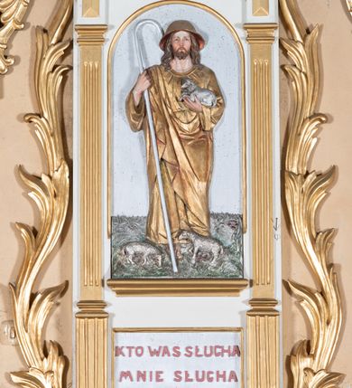 Zdjęcie nr 6: Ambona przyścienna, wisząca, z dostępem od lewej strony w postaci jednobiegowych schodów z balustradą. Kosz na planie sześcioboku na profilowanym cokole i ćwierćwałku; u spodu podwieszony profilowany ostrosłup o ściętym szczycie, zakończony szyszką. Ścianki kosza wydzielone kolumienkami  o trzonach dekorowanych do 1/3 wysokości kanelowaniem, o kapitelach korynckich, na cokołach zdobionych liśćmi akantu, dźwigającymi imposty oraz wspólny gzyms stanowiąc parapet. W polach ścianek płyciny z płaskorzeźbionymi półpostaciami czterech ewangelistów, każdy z nich ukazany został z złotej sukni, z identyfikującym go atrybutem, księgą lub zwojem i piórem w ręce –  od lewej św. Jan, św. Mateusz, św. Łukasz i św. Marek. Zaplecek w formie wydłużonego prostokąta, ujętego pilastrami podtrzymującymi imposty i wspólny gzyms. W polu płycina w formie stojącego prostokąta zamkniętego łukiem półkolistym z płaskorzeźbionym przedstawieniem Chrystusa Dobrego Pasterza, u spodu płycina z napisem „KTO WAS SŁUCHA / MNIE SŁUCHA”.  Chrystus został ukazany w całej postaci, lewą ręką podtrzymując baranka, a prawą laskę pasterską. Ma podłużna, poważną twarz, małe, niebieskie oczy, wąski nos i usta, broda dzieli się w dwa pukle; falowane włosy opadają na ramiona. Jest ubrany w długą, złotą tunikę i złoty płaszcz, na głowie ma złoty kalepusz, czerwony od wewnątrz. Stoi na trawie, u jego stóp dwa srebrzone baranki Zaplecek ujęty uszami w formie falujących liści. Baldachim powtarza kształt kosza, nakryty kopułką pokrytą łuską, z podwieszonym fryzem roślinnym, dekorowany w narożach palmetami. Baldachim stanowi podstawę dla latarni opartej na sześciu spływach wolutowych dekorowanych kampanulami, podtrzymujących belkowanie i kopułkę, stanowiącą cokół dla figury św. Jana Chrzciciela, który stoi w kontrapoście, prawą ręką unosi na wysokość twarzy muszlę, a e lewej trzyma trzyż. Ma podł€żną twarz okoczoną gęstym zarostem, wzniesiony ku górze wzrok, wąski nos i małe usta. Jest ubrany w tunikę ze srerzonego fuyta i złoty płaszcz wywiniety na prawym biodrze i przechodzący przez lewy bark. Wewnątrz latarni figura anioła na postumencie. Został ukazany w całej postaci, z rękami złożonymi na piersi. Ma okrągłą twarz o deliatnych rysach i falowane włosy opadające na kark. Jest ubrany w długa, złotą suknię w szerokomi rękawami. Z pleców wyrastają mu duże, srebrne skrzydła z ptacich piór. Na podniebiu baldachimu gołębica Ducha Świętego w glorii promienistej. Struktura polichromowana na biało, profile na czerwono, detale i ornamentyka złocone.