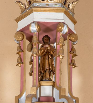 Zdjęcie nr 4: Ambona przyścienna, wisząca, z dostępem od lewej strony w postaci jednobiegowych schodów z balustradą. Kosz na planie sześcioboku na profilowanym cokole i ćwierćwałku; u spodu podwieszony profilowany ostrosłup o ściętym szczycie, zakończony szyszką. Ścianki kosza wydzielone kolumienkami  o trzonach dekorowanych do 1/3 wysokości kanelowaniem, o kapitelach korynckich, na cokołach zdobionych liśćmi akantu, dźwigającymi imposty oraz wspólny gzyms stanowiąc parapet. W polach ścianek płyciny z płaskorzeźbionymi półpostaciami czterech ewangelistów, każdy z nich ukazany został z złotej sukni, z identyfikującym go atrybutem, księgą lub zwojem i piórem w ręce –  od lewej św. Jan, św. Mateusz, św. Łukasz i św. Marek. Zaplecek w formie wydłużonego prostokąta, ujętego pilastrami podtrzymującymi imposty i wspólny gzyms. W polu płycina w formie stojącego prostokąta zamkniętego łukiem półkolistym z płaskorzeźbionym przedstawieniem Chrystusa Dobrego Pasterza, u spodu płycina z napisem „KTO WAS SŁUCHA / MNIE SŁUCHA”.  Chrystus został ukazany w całej postaci, lewą ręką podtrzymując baranka, a prawą laskę pasterską. Ma podłużna, poważną twarz, małe, niebieskie oczy, wąski nos i usta, broda dzieli się w dwa pukle; falowane włosy opadają na ramiona. Jest ubrany w długą, złotą tunikę i złoty płaszcz, na głowie ma złoty kalepusz, czerwony od wewnątrz. Stoi na trawie, u jego stóp dwa srebrzone baranki Zaplecek ujęty uszami w formie falujących liści. Baldachim powtarza kształt kosza, nakryty kopułką pokrytą łuską, z podwieszonym fryzem roślinnym, dekorowany w narożach palmetami. Baldachim stanowi podstawę dla latarni opartej na sześciu spływach wolutowych dekorowanych kampanulami, podtrzymujących belkowanie i kopułkę, stanowiącą cokół dla figury św. Jana Chrzciciela, który stoi w kontrapoście, prawą ręką unosi na wysokość twarzy muszlę, a e lewej trzyma trzyż. Ma podł€żną twarz okoczoną gęstym zarostem, wzniesiony ku górze wzrok, wąski nos i małe usta. Jest ubrany w tunikę ze srerzonego fuyta i złoty płaszcz wywiniety na prawym biodrze i przechodzący przez lewy bark. Wewnątrz latarni figura anioła na postumencie. Został ukazany w całej postaci, z rękami złożonymi na piersi. Ma okrągłą twarz o deliatnych rysach i falowane włosy opadające na kark. Jest ubrany w długa, złotą suknię w szerokomi rękawami. Z pleców wyrastają mu duże, srebrne skrzydła z ptacich piór. Na podniebiu baldachimu gołębica Ducha Świętego w glorii promienistej. Struktura polichromowana na biało, profile na czerwono, detale i ornamentyka złocone.