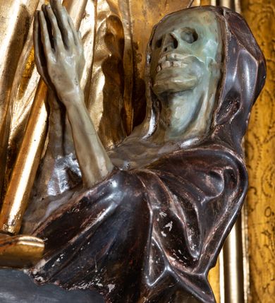 Zdjęcie nr 6: Rzeźba ścięta z tyłu, drążona, na cokole. Figura ustawiona frontalnie, w kontrapoście, z delikatnie pochyloną głową. W lewej ręce trzyma pastorał, prawą ma opuszczona, przed sobą. Twarz pociągła, o starczych rysach, z widocznymi zmarszczkami, oczy półprzymknięte, spoglądające w dół, nos prosty, usta pełne. Włosy zwinięte w pukle, głęboko. Święty ubrany w albę zapinaną na guziki, rokietę z rytą dekoracją roślinną u spodu, przepasane w talii; tkanina przylegająca do ciała, drapowana w liczne, drobne fałdki. Na piersi krzyż pektoralny i dystynktorium kanonickie. Kapa zapięta dekoracyjną fibulą, opadająca na plecy i ramiona, z kameryzowaną lamówką; tkanina sztywna, delikatnie drapowana. Na rękach czerwone rękawiczki, na głowie infuła. U lewej nogi świętego półpostać Piotrowina zwróconego profilem w lewo, ukazanego jako kościotrup, z narzuconym na czaszkę i ramiona fioletowym całunem. Polichromia naturalistyczna w odsłoniętych partiach ciała, szaty złocone, detal polichromowany.