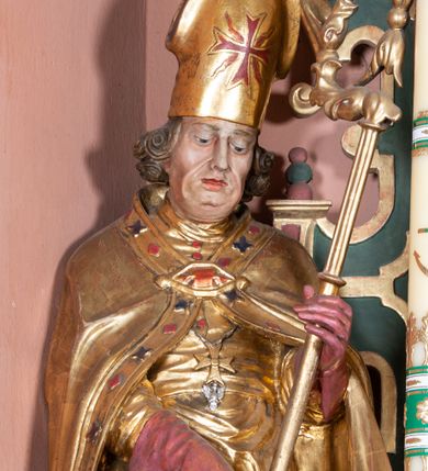 Zdjęcie nr 1: Rzeźba ścięta z tyłu, drążona, na cokole. Figura ustawiona frontalnie, w kontrapoście, z delikatnie pochyloną głową. W lewej ręce trzyma pastorał, prawą ma opuszczona, przed sobą. Twarz pociągła, o starczych rysach, z widocznymi zmarszczkami, oczy półprzymknięte, spoglądające w dół, nos prosty, usta pełne. Włosy zwinięte w pukle, głęboko. Święty ubrany w albę zapinaną na guziki, rokietę z rytą dekoracją roślinną u spodu, przepasane w talii; tkanina przylegająca do ciała, drapowana w liczne, drobne fałdki. Na piersi krzyż pektoralny i dystynktorium kanonickie. Kapa zapięta dekoracyjną fibulą, opadająca na plecy i ramiona, z kameryzowaną lamówką; tkanina sztywna, delikatnie drapowana. Na rękach czerwone rękawiczki, na głowie infuła. U lewej nogi świętego półpostać Piotrowina zwróconego profilem w lewo, ukazanego jako kościotrup, z narzuconym na czaszkę i ramiona fioletowym całunem. Polichromia naturalistyczna w odsłoniętych partiach ciała, szaty złocone, detal polichromowany.