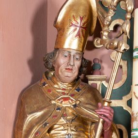 Zdjęcie nr 1: Rzeźba ścięta z tyłu, drążona, na cokole. Figura ustawiona frontalnie, w kontrapoście, z delikatnie pochyloną głową. W lewej ręce trzyma pastorał, prawą ma opuszczona, przed sobą. Twarz pociągła, o starczych rysach, z widocznymi zmarszczkami, oczy półprzymknięte, spoglądające w dół, nos prosty, usta pełne. Włosy zwinięte w pukle, głęboko. Święty ubrany w albę zapinaną na guziki, rokietę z rytą dekoracją roślinną u spodu, przepasane w talii; tkanina przylegająca do ciała, drapowana w liczne, drobne fałdki. Na piersi krzyż pektoralny i dystynktorium kanonickie. Kapa zapięta dekoracyjną fibulą, opadająca na plecy i ramiona, z kameryzowaną lamówką; tkanina sztywna, delikatnie drapowana. Na rękach czerwone rękawiczki, na głowie infuła. U lewej nogi świętego półpostać Piotrowina zwróconego profilem w lewo, ukazanego jako kościotrup, z narzuconym na czaszkę i ramiona fioletowym całunem. Polichromia naturalistyczna w odsłoniętych partiach ciała, szaty złocone, detal polichromowany.