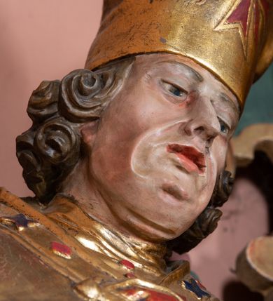 Zdjęcie nr 4: Rzeźba ścięta z tyłu, drążona, na cokole. Figura ustawiona frontalnie, w kontrapoście, z delikatnie pochyloną głową. W lewej ręce trzyma pastorał, prawą ma opuszczona, przed sobą. Twarz pociągła, o starczych rysach, z widocznymi zmarszczkami, oczy półprzymknięte, spoglądające w dół, nos prosty, usta pełne. Włosy zwinięte w pukle, głęboko. Święty ubrany w albę zapinaną na guziki, rokietę z rytą dekoracją roślinną u spodu, przepasane w talii; tkanina przylegająca do ciała, drapowana w liczne, drobne fałdki. Na piersi krzyż pektoralny i dystynktorium kanonickie. Kapa zapięta dekoracyjną fibulą, opadająca na plecy i ramiona, z kameryzowaną lamówką; tkanina sztywna, delikatnie drapowana. Na rękach czerwone rękawiczki, na głowie infuła. U lewej nogi świętego półpostać Piotrowina zwróconego profilem w lewo, ukazanego jako kościotrup, z narzuconym na czaszkę i ramiona fioletowym całunem. Polichromia naturalistyczna w odsłoniętych partiach ciała, szaty złocone, detal polichromowany.