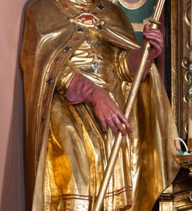 Zdjęcie nr 2: Rzeźba ścięta z tyłu, drążona, na cokole. Figura ustawiona frontalnie, w kontrapoście, z delikatnie pochyloną głową. W lewej ręce trzyma pastorał, prawą ma opuszczona, przed sobą. Twarz pociągła, o starczych rysach, z widocznymi zmarszczkami, oczy półprzymknięte, spoglądające w dół, nos prosty, usta pełne. Włosy zwinięte w pukle, głęboko. Święty ubrany w albę zapinaną na guziki, rokietę z rytą dekoracją roślinną u spodu, przepasane w talii; tkanina przylegająca do ciała, drapowana w liczne, drobne fałdki. Na piersi krzyż pektoralny i dystynktorium kanonickie. Kapa zapięta dekoracyjną fibulą, opadająca na plecy i ramiona, z kameryzowaną lamówką; tkanina sztywna, delikatnie drapowana. Na rękach czerwone rękawiczki, na głowie infuła. U lewej nogi świętego półpostać Piotrowina zwróconego profilem w lewo, ukazanego jako kościotrup, z narzuconym na czaszkę i ramiona fioletowym całunem. Polichromia naturalistyczna w odsłoniętych partiach ciała, szaty złocone, detal polichromowany.