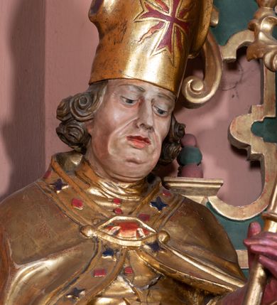 Zdjęcie nr 3: Rzeźba ścięta z tyłu, drążona, na cokole. Figura ustawiona frontalnie, w kontrapoście, z delikatnie pochyloną głową. W lewej ręce trzyma pastorał, prawą ma opuszczona, przed sobą. Twarz pociągła, o starczych rysach, z widocznymi zmarszczkami, oczy półprzymknięte, spoglądające w dół, nos prosty, usta pełne. Włosy zwinięte w pukle, głęboko. Święty ubrany w albę zapinaną na guziki, rokietę z rytą dekoracją roślinną u spodu, przepasane w talii; tkanina przylegająca do ciała, drapowana w liczne, drobne fałdki. Na piersi krzyż pektoralny i dystynktorium kanonickie. Kapa zapięta dekoracyjną fibulą, opadająca na plecy i ramiona, z kameryzowaną lamówką; tkanina sztywna, delikatnie drapowana. Na rękach czerwone rękawiczki, na głowie infuła. U lewej nogi świętego półpostać Piotrowina zwróconego profilem w lewo, ukazanego jako kościotrup, z narzuconym na czaszkę i ramiona fioletowym całunem. Polichromia naturalistyczna w odsłoniętych partiach ciała, szaty złocone, detal polichromowany.