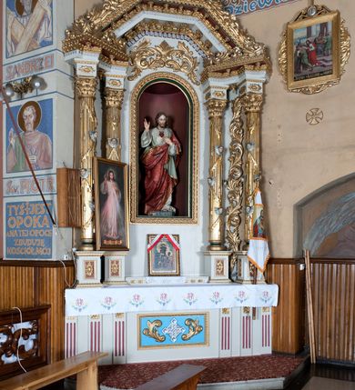 Zdjęcie nr 4: Nastawa ołtarzowa architektoniczna, jednoosiowa, jednokondygnacyjna ze zwieńczeniem. Ołtarz drewniany, prostopadłościenny. Mensa prostopadłościenna, z antepedium dekorowanym płyciną w kształcie leżącego prostokąta z krzyżem greckim o ramionach zakończonych rombami, na tle rombu i ujętym liśćmi. Flankują ją z każdej strony trzy pilastry o żłobkowanych trzonach. Na mensie, na cokole obrazem z przedstawieniem Matki Boskiej Częstochowskiejj. Nastawa ujęta parami kolumn na cokołach, z których zewnętrzne są wysunięte. Podpory dźwigają pełne belkowanie, którego architraw i fryz tworzą zygzak, w gzyms jest prosty. Na nich wspierają się trzy równoległe, załamane przyczółki zakomponowane uskokowo. Szczyt tworzy krzyż łaciński stylizowany na kwiat osadzony na łodydze z liśćmi. Pole środkowe ujęte profilowaną ramą z półokrągłą niszą wypełnioną figurą Najświętszego Serca Jezusa. Cokoły kolumn dekorowane płycinami z plakietami roślinnymi, trzony kolumn uskrzydlonymi główkami anielskimi podtrzymującymi festony utworzone z tkaniny i chwosty, między kolumnami ażurowa wić akantowa. Belkowanie ozdabia kostkowanie i astragal, pole nad obrazem wypełnia symetryczna wić akantowa, która wieńczy również gzyms belkowania i zewnętrzną krawędź przyczółków. Gzyms od spodu dekorowany rozetami. Struktura polichromowana na biało i jasnoszaro, kolumny, ornamenty i dekoracje złocone, płyciny czerwone i niebieskie; fragmenty baz kolumn zielone; nisza niebieska. 