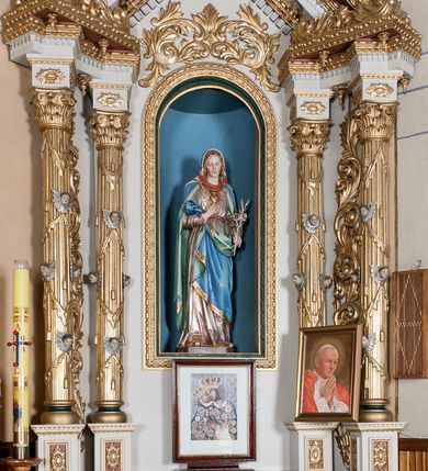 Zdjęcie nr 2: Nastawa ołtarzowa architektoniczna, jednoosiowa, jednokondygnacyjna ze zwieńczeniem. Ołtarz drewniany, prostopadłościenny. Mensa prostopadłościenna, z antepedium dekorowanym płyciną w kształcie leżącego prostokąta z krzyżem greckim o ramionach zakończonych rombami, na tle rombu i ujętym liśćmi. Flankują ją z każdej strony trzy pilastry o żłobkowanych trzonach. Na mensie, na cokole obraz z przedstawieniem Matki Boskiej Kalwaryjskiej. Nastawa ujęta parami kolumn na cokołach, z których zewnętrzne są wysunięte. Podpory dźwigają pełne belkowanie, którego architraw i fryz tworzą zygzak, a gzyms jest prosty. Na nich wspierają się trzy równoległe, załamane przyczółki zakomponowane uskokowo. Szczyt tworzy krzyż łaciński stylizowany na kwiat osadzony na łodydze z liśćmi. Pole środkowe ujęte profilowaną ramą z półokrągłą niszą wypełnioną figurą Najświętszego Serca Marii. Cokoły kolumn dekorowane płycinami z plakietami roślinnymi, trzony kolumn uskrzydlonymi główkami anielskimi podtrzymującymi festony utworzone z tkaniny i chwosty, między kolumnami ażurowa wić akantowa. Belkowanie ozdabia kostkowanie i astragal, pole nad obrazem wypełnia symetryczna wić akantowa, która wieńczy również gzyms belkowania i zewnętrzną krawędź przyczółków. Gzyms od spodu dekorowany rozetami. Struktura polichromowana na biało i jasnoszaro, kolumny, ornamenty i dekoracje złocone, płyciny czerwone i niebieskie; fragmenty baz kolumn zielone; nisza niebieska. 