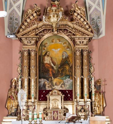 Zdjęcie nr 6: Nastawa ołtarzowa architektoniczna, jednoosiowa, jednokondygnacyjna ze zwieńczeniem. Ołtarz drewniany, prostopadłościenny. Mensa drewniana, o silnie wysuniętym gzymsie dekorowanego kostkowaniem. Antepedium w formie leżącego prostokąta z płyciną o dwóch wypukłych bokach. W polu płaskorzeźbione przedstawienie pelikana karmiącego w gnieździe młode, flankowanego winnym gronem i kłosami zbóż; płycina ujęta parą uskrzydlonych główek anielskich. Antepedium zamknięte profilowanym gzymsem, dekorowanym kostkowaniem, dźwigającym niepełne belkowanie. Tabernakulum prostopadłościenne, w formie drewnianej skrzynki obitej blachą miedzianą, ustawione na wysokim cokole ujętym od frontu i po bokach spływami wolutowymi, dźwigającymi wyłamany gzyms dekorowany kostkowaniem; na froncie płycina z rozetą. Drzwiczki w kształcie leżącego prostokąta zamkniętego łukiem segmentowym z aplikowanym kielichem z hostią, w otoczeniu winnego grona, na fakturowanym tle. Tabernakulum flankowane parą kanelowanych kolumienek, dźwigających gzyms wieńczący, dekorowany kostkowaniem oraz szczyt w formie zbliżonej do trójkąta, zamknięty gzymsem przechodzącym w przeciwstawnie zwrócone woluty, dekorowane kostkowaniem; w polu uskrzydlona główka anielska. 
Retabulum na zryzalitowanym przy brzegach cokole; ujęte dwiema parami kolumn o kapitelach kompozytowych i kanelowanych trzonach dźwigających pełne belkowanie oraz trójkątny przyczółek trójbocznie załamany; na nim, na osi podpór przerwany, wolutowy przyczółek, z leżącymi nań figurami. Szczyt w postaci dwustrefowego cokołu ujętego kartuszem z figurą św. Michała Archanioła. Pole środkowe w całości wypełnia płytka nisza ujęta profilowaną ramą z obrazem „Trójca Święta”, wymiennie z przedstawieniami Matki Boskiej z Dzieciątkiem i św. Katarzyny. Kondygnację flankują, ustawione na cokołach figury św. Stanisława i św. Wojciecha. Cokoły kolumn dekorowane płycinami z uskrzydlonymi główkami anielskimi; w polu cokołu płycina w formie wydłużonego, leżącego prostokąta o wypukłych bokach, z dekoracją schweifwerkową. Trzony kolumn dekorowane uskrzydlonymi główkami anielskimi podtrzymującymi festony utworzone z tkaniny i chwosty. Za kolumnami płyciny wypełnione schweifwerkiem, w ich centrum obelisk dekorowany cekinami i zwieńczony krzyżem. W partii fryzu od frontu uskrzydlone główki anielskie, po bokach schweifwerk. Pola obu przyczółków wypełnia ornament schweifwerkowy. Gzyms od spodu dekorowany rozetami; zwieńczony wolutowym kluczem dekorowanym schwrifwerkiem oraz uskrzydloną główką anielską. Woluty wieńczące zdobione palmetami. Kondygnacja ujęta uszami utworzonymi z ornamentu okuciowego i ulistnionych wolut. Struktura w całości złocona, płyciny i profile polichromowane na czerwono, uszaki na zielono. 