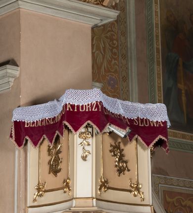 Zdjęcie nr 1: Ambona przyścienna wisząca, z dostępem od strony prezbiterium w postaci dwubiegowych schodów z podestem. Balustrada pełna, na cokole, dekorowana płycinami w formie rombów, z poręczą w postaci gzymsu. Kosz na planie kwadratu o ściętych narożach i wklęsłych bokach, na cokole w formie spłaszczonego wałka dekorowanego kampanulami w układzie poziomym. U spodu podwieszony odwrócony ostrosłup o ściętym wierzchołku i wklęsłych bokach ujęty w narożach spływami wolutowymi, dekorowany kampanulami, zakończony szyszką zdobioną cęgami. Ścianki kosza wydzielone pilastrami w formie wolut, których trzony dekorowane są ukwieconymi gałązkami. Na ściankach rocaille'owe płyciny z ornamentem roślinnym. Parapet w postaci gzymsu. Baldachim na planie analogicznym względem kosza, w formie gzymsu, dekorowany stylizowanym perełkowaniem, z podwieszonym lambrekinem. Na nim, na podstawie utworzonej z obłoków figura Chrystusa, zwróconego frontalnie, trzymającego na lewym ramieniu krzyż, z prawą ręką wskazującą w górę. Po jego lewej figurka Mojżesza z tablicami przykazań, po prawej św. Michała Archanioła trzymającego trąbę i wagę. Na podniebieniu baldachimu płaskorzeźbiona gołębica Ducha Świętego z czterema wiązkami promieni oraz dwie ukwiecone gałązki. Struktura polichromowana w kolorze beżowym, z białym detalem, ornamentyka i profile złocone, obłoki i gołębica srebrzone.