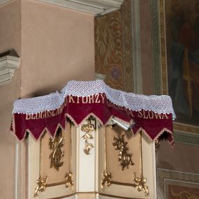Zdjęcie nr 1: Ambona przyścienna wisząca, z dostępem od strony prezbiterium w postaci dwubiegowych schodów z podestem. Balustrada pełna, na cokole, dekorowana płycinami w formie rombów, z poręczą w postaci gzymsu. Kosz na planie kwadratu o ściętych narożach i wklęsłych bokach, na cokole w formie spłaszczonego wałka dekorowanego kampanulami w układzie poziomym. U spodu podwieszony odwrócony ostrosłup o ściętym wierzchołku i wklęsłych bokach ujęty w narożach spływami wolutowymi, dekorowany kampanulami, zakończony szyszką zdobioną cęgami. Ścianki kosza wydzielone pilastrami w formie wolut, których trzony dekorowane są ukwieconymi gałązkami. Na ściankach rocaille'owe płyciny z ornamentem roślinnym. Parapet w postaci gzymsu. Baldachim na planie analogicznym względem kosza, w formie gzymsu, dekorowany stylizowanym perełkowaniem, z podwieszonym lambrekinem. Na nim, na podstawie utworzonej z obłoków figura Chrystusa, zwróconego frontalnie, trzymającego na lewym ramieniu krzyż, z prawą ręką wskazującą w górę. Po jego lewej figurka Mojżesza z tablicami przykazań, po prawej św. Michała Archanioła trzymającego trąbę i wagę. Na podniebieniu baldachimu płaskorzeźbiona gołębica Ducha Świętego z czterema wiązkami promieni oraz dwie ukwiecone gałązki. Struktura polichromowana w kolorze beżowym, z białym detalem, ornamentyka i profile złocone, obłoki i gołębica srebrzone.